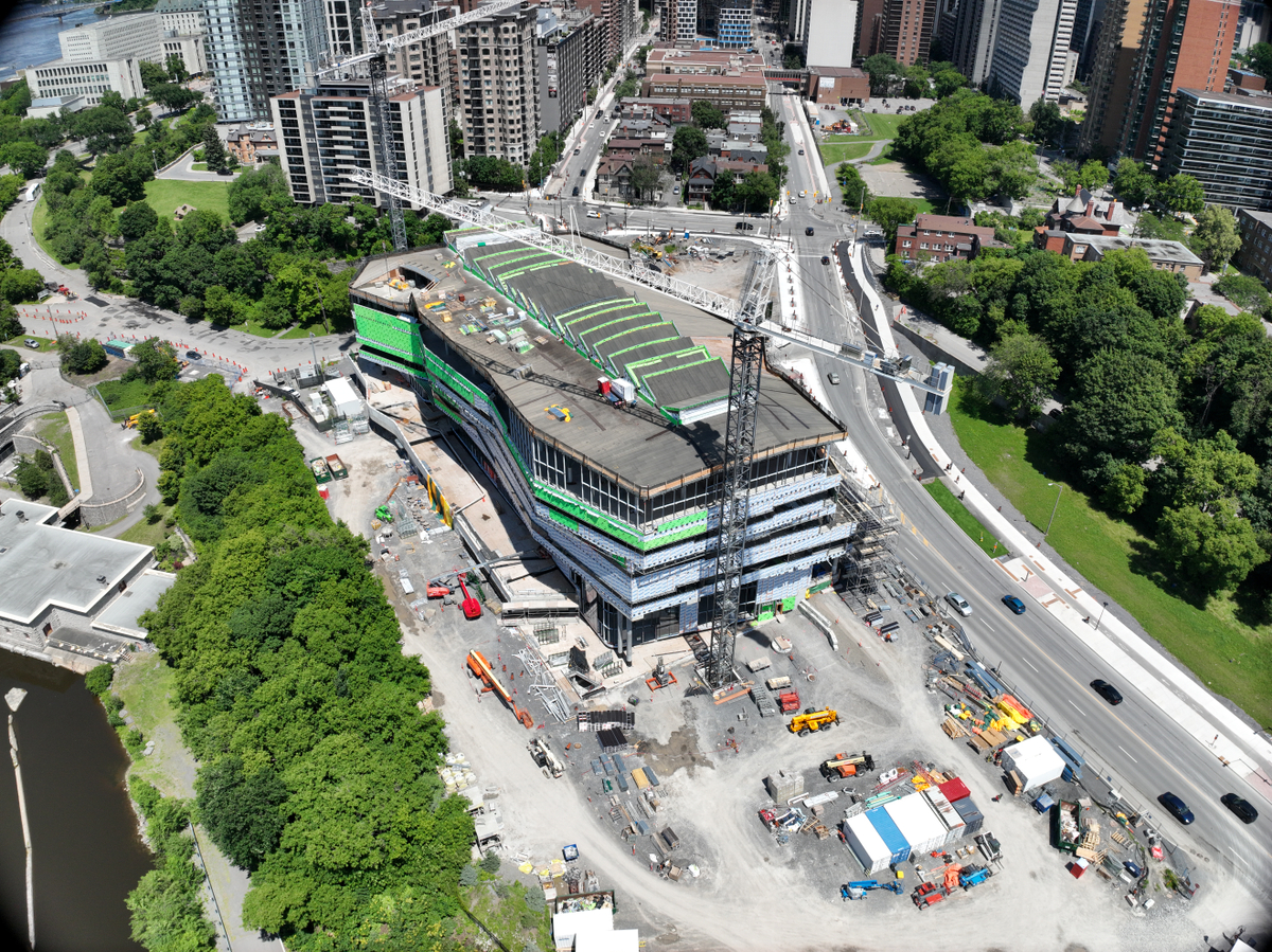 Aerial view of the Ādisōke facility showing the wooden roof paneling, with a tower crane overhead. Several tall buildings are visible in the background.