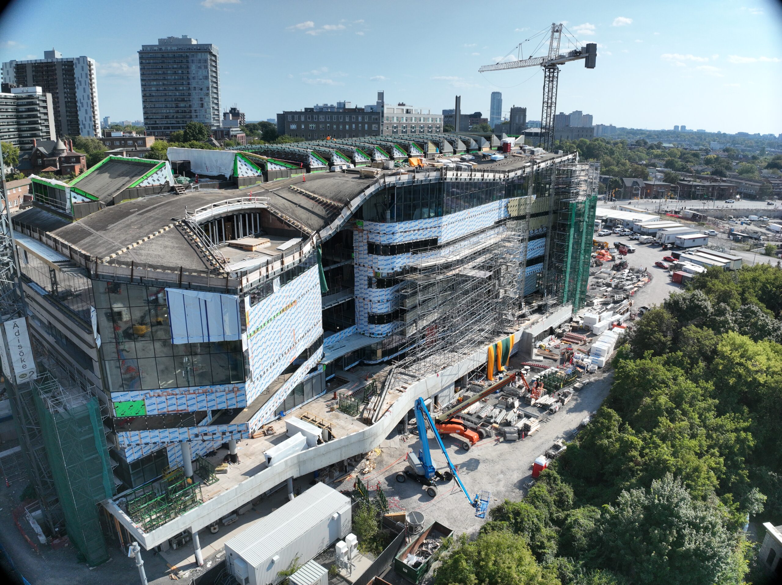 Photo prise par un drone de l’installation Ādisōke; on y voit les lignes incurvées de la toiture en bois. À l’arrière-plan, on aperçoit des immeubles de grande hauteur et une grue de chantier.