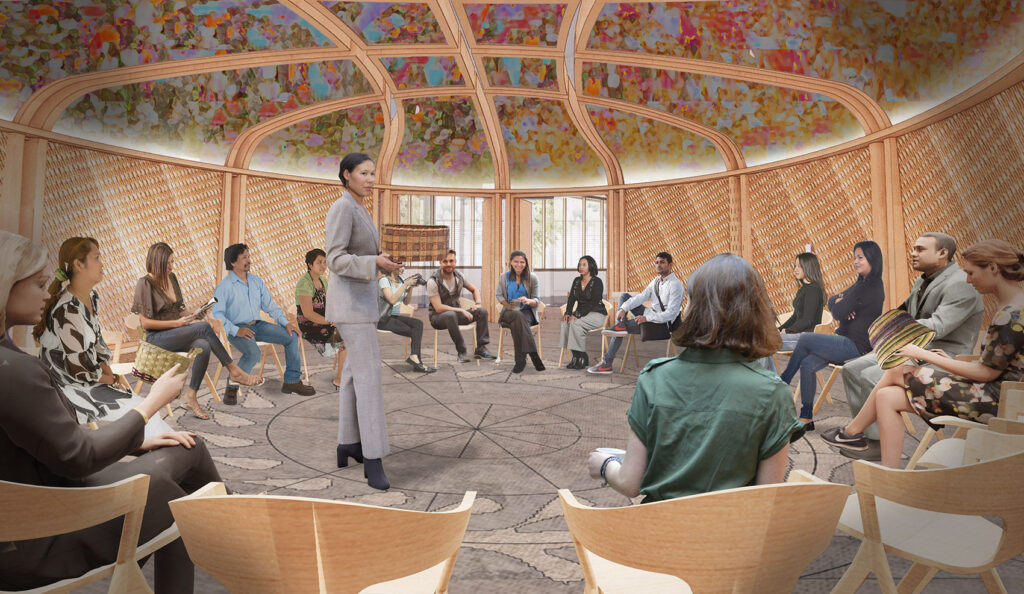 Group seated in a circle inside an indigenous circular lodge with wooden walls and a colorful ceiling; a person of indigenous origin stands in the center, addressing the group.